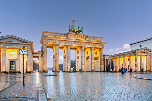 Brandenburg Gate, a neoclassical monument with 12 Doric columns and a chariot sculpture on top, illuminated by lights. The image is taken from the Pariser Platz, a square in front of the gate, where the ground is wet from rain. The sky is blue with clouds.