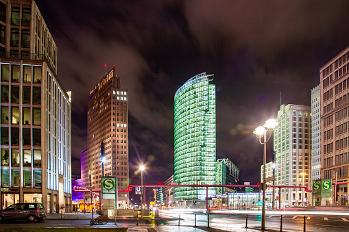 A city street at night in Berlin Potsdamer Platz, a public square and traffic intersection. The image is taken from a low angle, looking up at the tall buildings and bright lights. The buildings are modern and have a lot of glass windows. The building on the right is curved and has a greenish hue. The building on the left is rectangular and has a red hue. The street is wet and there are reflections of the lights on the pavement. There are street lamps and traffic lights visible. There is a black car on the left side of the street.
