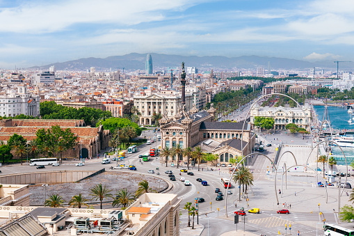 A panoramic view of Barcelona, Spain. The image is taken from a high vantage point, looking down on the city. The image shows the city’s dense urban landscape, with many buildings and streets visible. The image also shows the city’s famous landmarks, such as the Columbus Monument, a tall column honoring the explorer. The image shows the city’s coastline, with the Mediterranean Sea visible in the background. The image shows a busy street with cars and people visible.