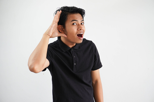 young Asian man looking camera and doing can not hear gesture, trying to listening a gossip wearing black polo t shirt isolated on white background