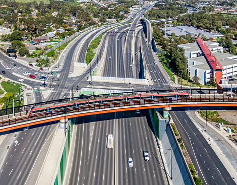 Aerial view modern electric train travelling on bridge over wide, multi-lane highway with elevated intersection, interchange and overpasses in treelined suburb. Flinders Rail Link, Southern Expressway, Main South Road, Sturt Road and Science Park all visible with Darlington in background. Logos & ID edited.