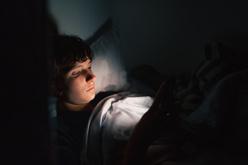 teenager uses gadget in dark. Teen girl, lying under covers in bed, uses mobile phone, at night