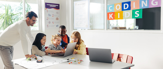 Primary school children learn to program robots with their teacher's guidance in a classroom. Group of kids using a laptop to develop a code that can control robotic cars in a STEM lesson.