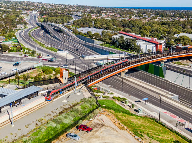 Aerial view modern electric train on bridge over multi-lane highway with intersection, interchange in treelined suburb with ocean view