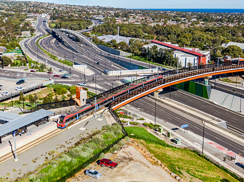 Essential & efficient rail infrastructure: aerial view modern electric train entering terminus station after exiting bridge over wide, multi-lane highway with elevated intersection, interchange and overpasses in treelined suburb. Flinders Rail Link, Southern Expressway, Main South Road, Sturt Road and Science Park all visible with Darlington service centres and ocean in background. Logos & ID edited.