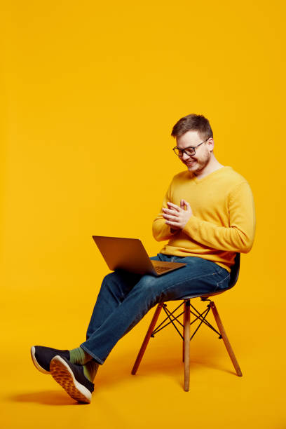 istock Excited young guy using laptop while sitting on chair, isolated over yellow background 1591985672