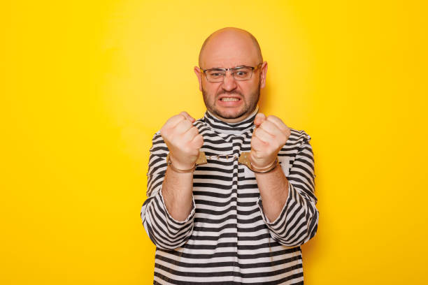 Man wearing prison uniform costume and handcuffs having fun making faces on yellow colored background with copy space