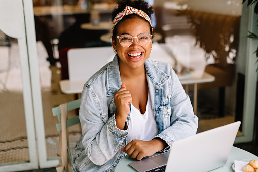 Young woman sits in a café, working on her laptop. She is a freelancer and content writer, enjoying the freedom of remote work. With a smile, she embodies the happiness of a successful entrepreneur.