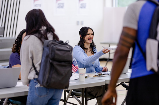 As several unrecognizable people are in line, the cheerful young adult female poll worker hands a paper ballot to an unseen voter.