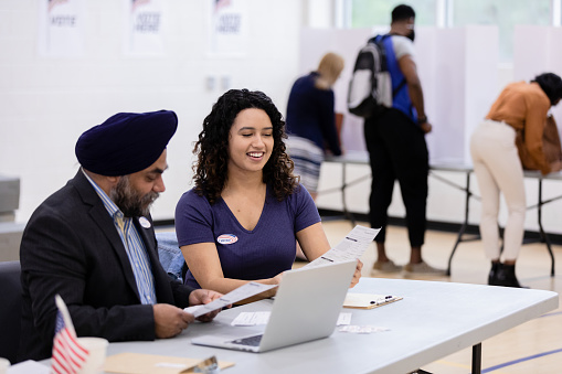 Two diverse election volunteers look at the ballot as people vote behind them.