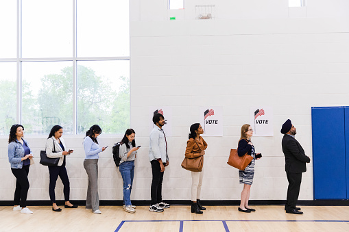 A multiracial group of people, many with smart phones, line up along a wall in a school gymnasium in order to vote.