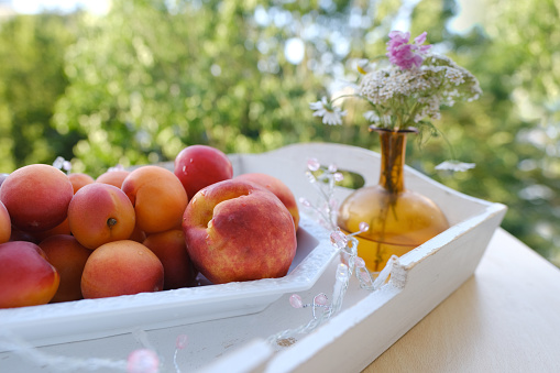 on wooden white tray plate with orange apricots, peach, bunch wild flowers on table in garden, beautiful summer still life, fruits, green trees in background, concept picnic in nature, healthy eating