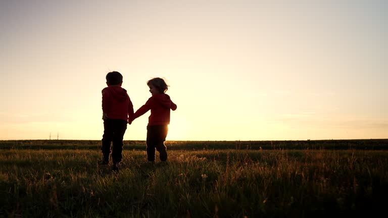 Silhouette of two little boys running together to the sun on open area field , friends holding hands. Children is our future, kids playing. Nature background. Happy childhood.