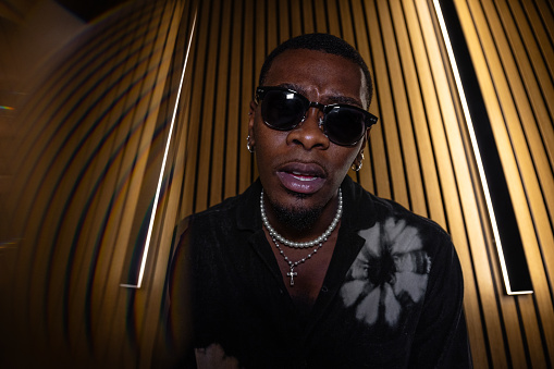 A low-angle portrait of a young black male wearing sunglasses in warm ambient lighting. He is looking down into the camera and is wearing pearls and a silver necklace around his neck. There is ambient lighting on both sides of him.