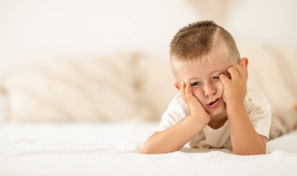 istock Unhappy tired little boy lying on bed and grimacing. 1592196316