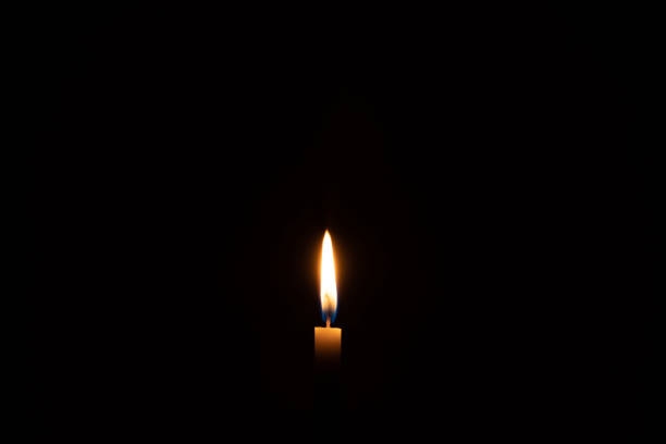 istock Single burning candle flame or light glowing on a small white candle on black or dark background on table in church for Christmas, funeral or memorial service with copy space. 1592231976