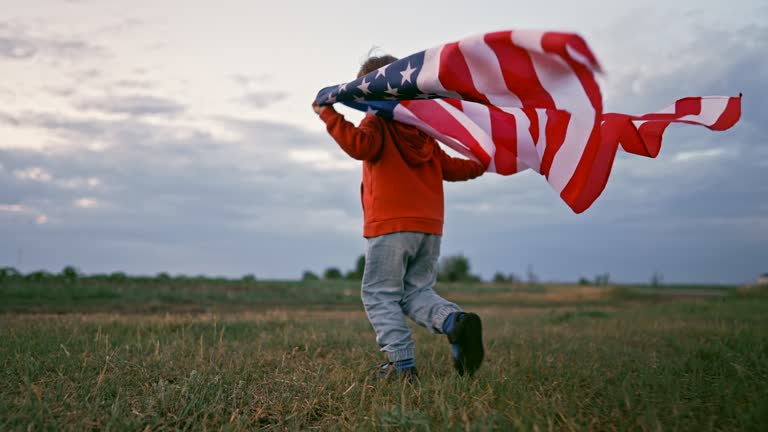 Cute little boy - American patriot kid running with national flag on open area field. USA, 4th of July - Independence day, celebration. US banner, memorial Veterans Day, election, America, labor.