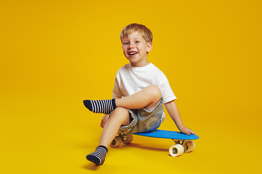 Cheerful blonde kid boy in white tshirt smiling and looking at camera while sitting on modern skateboard against yellow background