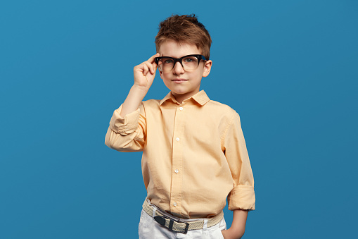 Little serious doubtful boy touching eyeglasses and looking at camera, standing isolated over blue background. Horizontal studio shot.