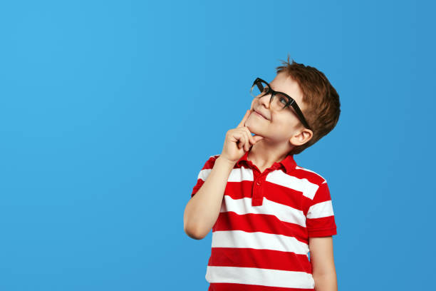 istock Smart kid in nerdy glasses and red striped shirt touching cheek and looking up while thinking 1592298016