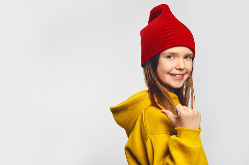 Good looking preteen girl wearing warm clothes, pointing with thumb on copy space for your advertising, has excited surprised happy expression, isolated on white background