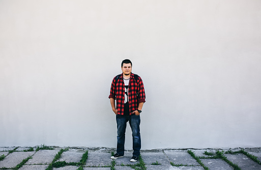 Young hipster guy in black and red checkered shirt is standing outdoors near the wall.