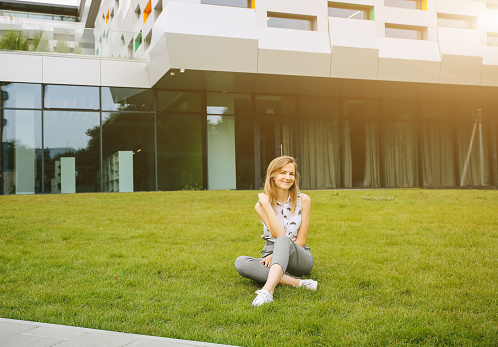 Beautiful hipster girl is sitting near the modern architecture building on the sunny day. Stylish business woman resting on the green grass lawn. Student's life free time. Office worker break.