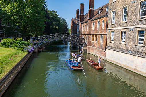 Cambridge UK 13th June 2023 \nPunting under the mathematical bridge over the River Cam in Cambridge \nSome punt boats with people sitting \ngreen foliage and purple flowers line the river bank