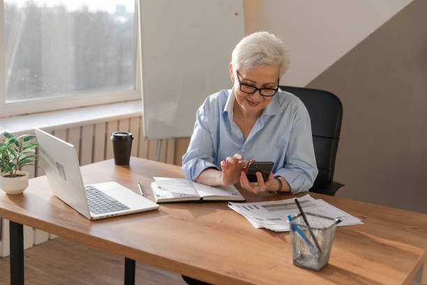 istock Confident stylish european middle aged senior woman using smartphone at workplace. Stylish older mature 60s gray haired lady businesswoman with cell phone in office. Boss leader using internet apps. 1592470708