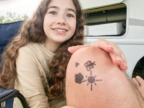 Daughter proudly showing drawings she made on father's knee with a permanent marker  during summer day besides motor home.