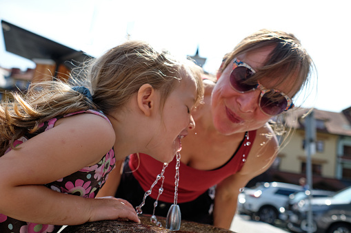 Beautiful mid adult mother is drinking water with her daughter at outdoor water fountain in summer