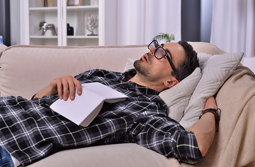 Bookish tranquility. A young man with facial hair wearing glasses fell asleep while reading a book on a plush couch in the inviting ambiance of his living room. Relaxation and literature concept.