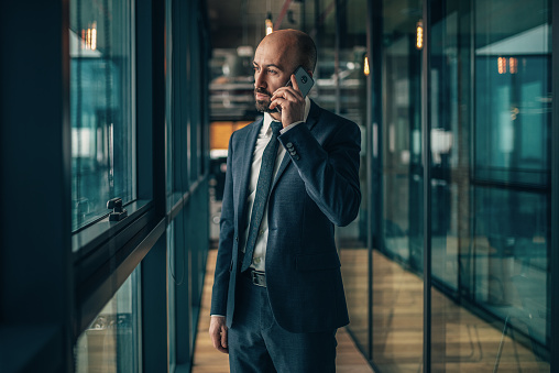 Portrait of Confident mid adult businessman standing in modern office and using smartphone. Elegant senior manager in office corridor talking to the phone