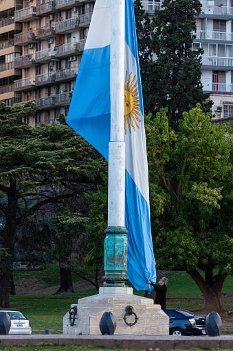 The big main flag of the Argentine National Flag Monument, being haul down at the afternoon, like each day. A person receives the flag. Behind, the National park and residentials buildings. On day: 1 August, 2023. Rosario city, Santa Fe province Argentina.