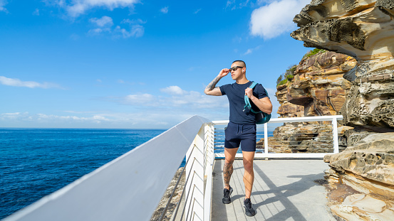 Asian man listening to the music on earphones and hiking on rocky mountain coastline in Sydney, Australia on summer vacation.