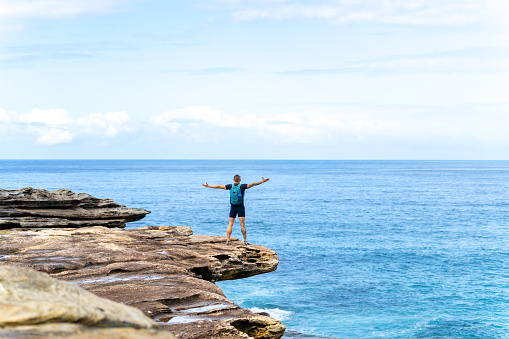 Rear view of Asian man standing on rocky mountain coastline with arm raised during travel at the beach in Sydney, Australia on summer vacation.