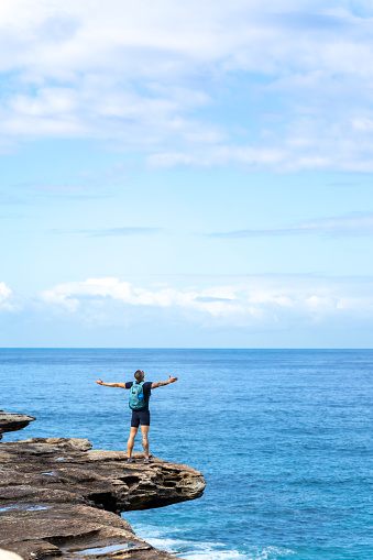 Rear view of Asian man standing on rocky mountain coastline with arm raised during travel at the beach in Sydney, Australia on summer vacation.