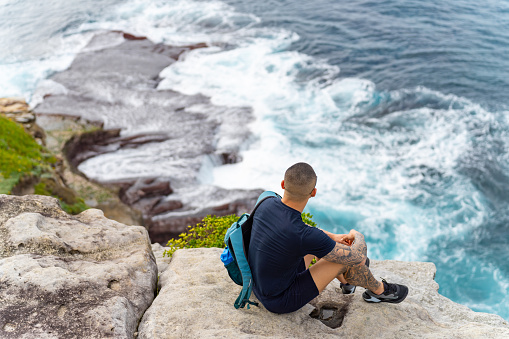 Asian man sitting on rocky mountain coastline during travel at the beach in Sydney, Australia on summer vacation.