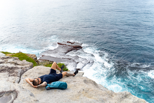 Asian man lying down and napping  on rocky mountain coastline during travel at the beach in Sydney, Australia on summer vacation.