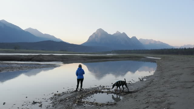 Woman walks dog by lakeshore, mountains distant