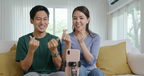 Young adult cute asia couple fun play dancing music game on IG reel tiktok instagram app at home selfie camera record video stories. Man woman youtuber joy showing vlog trendy talent sit relax at sofa joven adulto linda asia pareja divertida jugar juego de música de baile en ig reel tiktok instagram aplicación en casa selfie cámara grabar historias de video. hombre mujer youtuber alegría mostrando vlog talento de moda sentarse a relajarse en el sof�