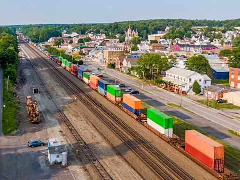 Aerial wide view vital modern rail infrastructure with heavy freight train carrying colourful container stacks passing through picturesque small rural Pennsylvania town in beautiful forested countryside. Three busy mainline tracks pass through the town providing essential transport links to sustain economic activity. Neat, well-maintained properties can be seen on the frontage street with church and spire in the background. In the foreground a spare track with stored railway equipment.  Logos and ID marks edited.