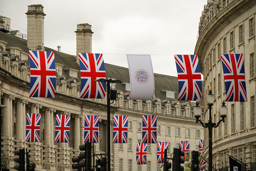 London, England, United Kingdom - May 7, 2023. Multiple British Union Jack flags with a King Charles III Coronation flag, a white flag with a royal monarch coronation seal, logo. The UK flags decorate Regent Street, sometimes called the Mile of Style, in London in honor of King Charles III and Queen Camilla's coronation. The red white and blue UK flags fill London streets during coronations, major events and holidays. The UK Union Jack flags bunting over Strand Street is a truly festive London city street scene with traditional English city buildings, architecture.