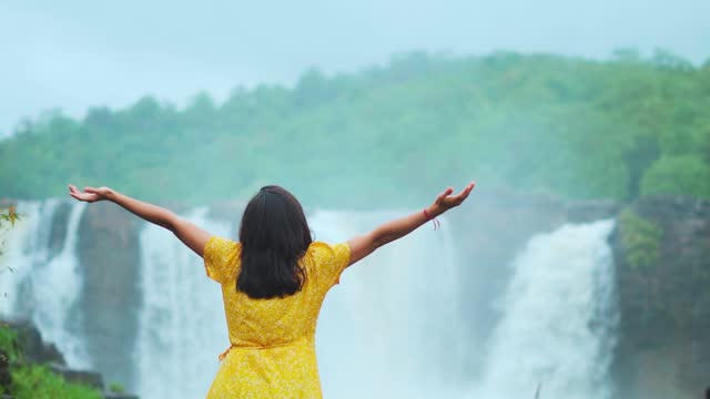 Tourist teenager girl spread arms in front of beautiful Gira waterfall, Gujarat, India. Female enjoying vacation on tourist destination. Waterfall flow during monsoon.