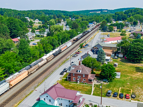 Aerial wide view vital modern rail infrastructure with two heavy freight trains passing through picturesque small rural Pennsylvania town in beautiful forested countryside. Three busy mainline tracks pass through the town providing essential transport links to sustain economic activity. Neat, well-maintained houses can be seen on both sides of the railroad tracks. Logos and ID marks edited.