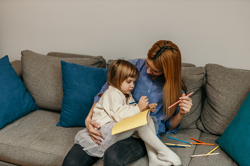 A mother and her young daughter sitting side by side on a cozy sofa in their living room.