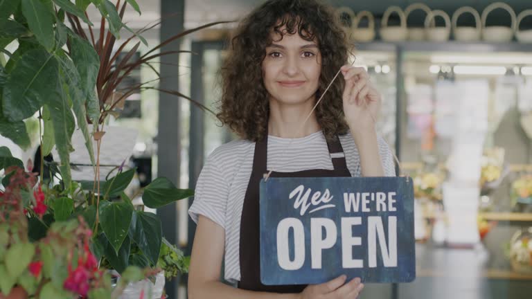 Portrait of young woman in apron changing Sorry we are closed to Yes we are open sign in flower shop welcoming customers