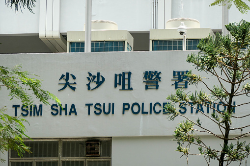 Facade of the Tsim Sha Tsui Police Station on Nathan Road, Kowloon, behind jacaranda and other tree branches and leaves.  Security bars are visible inside the window on the bottom left.  This image was taken on a sunny afternoon on 6 July 2023.