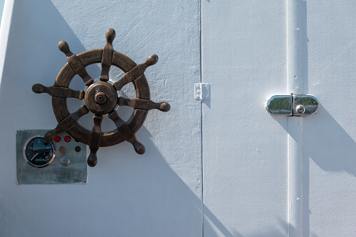 Close-up of the white metal wall of the cabin of a small sea boat. There is a wooden steering wheel, controls and a door lock. Background.