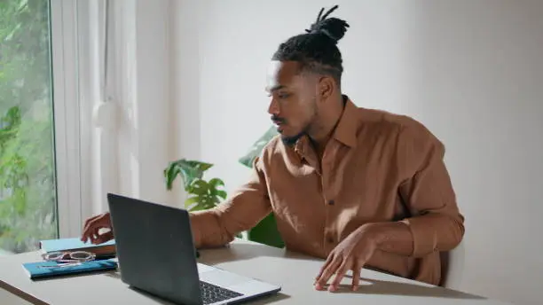 Focused man taking glasses on in office closeup. Dreadlocks guy working laptop at home remotely. Serious freelancer looking computer screen preparing taking notes. Afro american student sitting alone